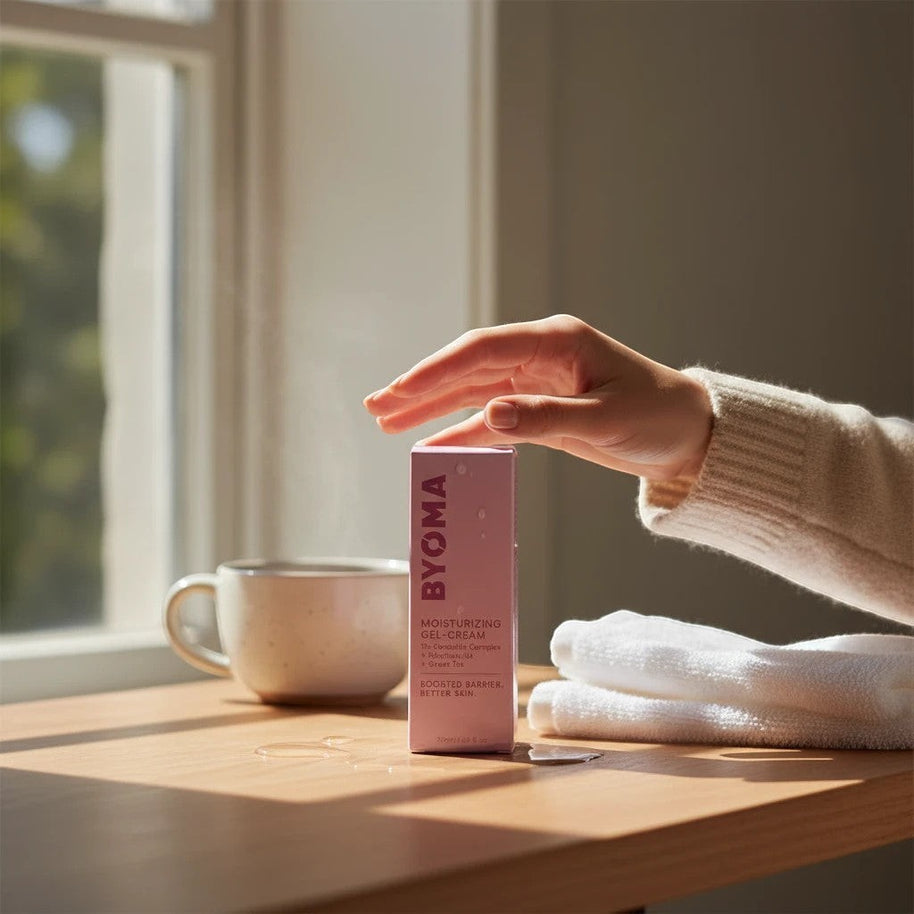 Pulse 360º Glow Byoma Moisturising Gel-Cream box held by a hand on a sunny wooden table with a mug and towels.